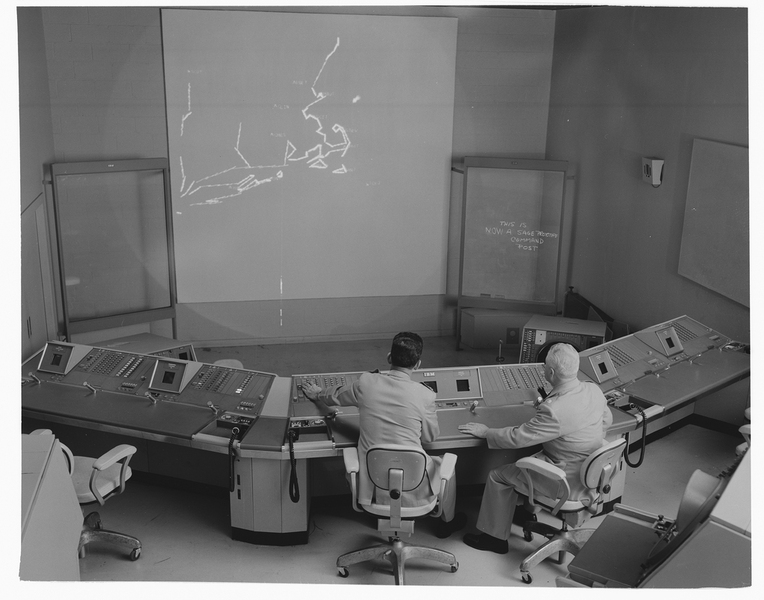 Grayscale photo of operators sitting in front of a digital display showing a crude line coastal map centered on Cape Cod