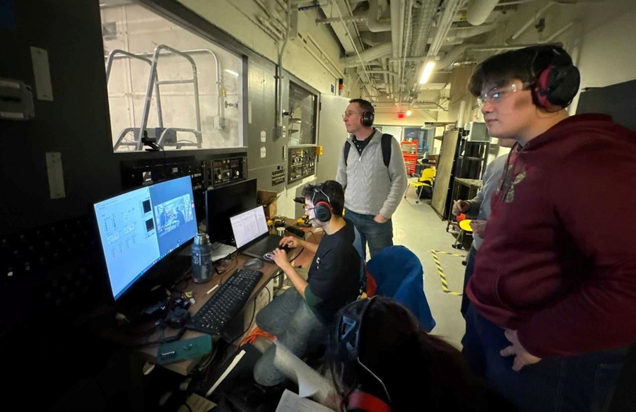 Four students wearing headphones watch their machinery being tested in a chamber (out of frame). One student sits at a computer desktop.