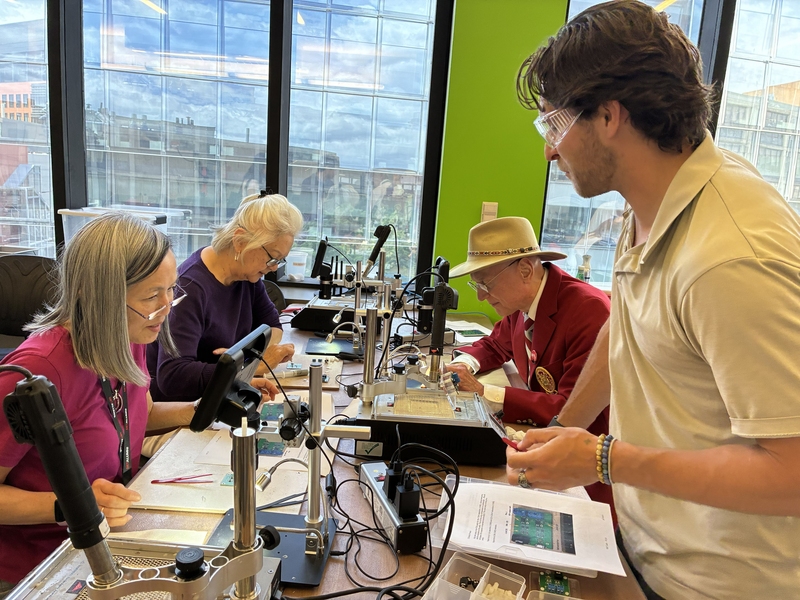 Staff member Liam Ackerman watches as three alumni practice surface mount soldering. 