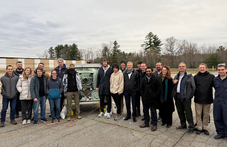 20 people flank a small jet engine in a parking lot on a cloudy winter day