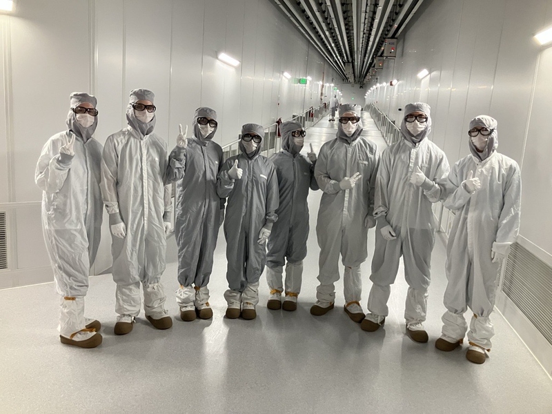 Group of 8 students in white lab suits and safety glasses standing in a hallway, making peace or thumbs-up hand gestures