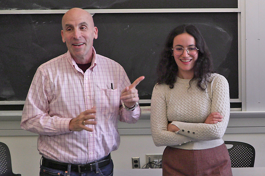 Ken Zolot gestures while standing next to a young woman in front of a blackboard