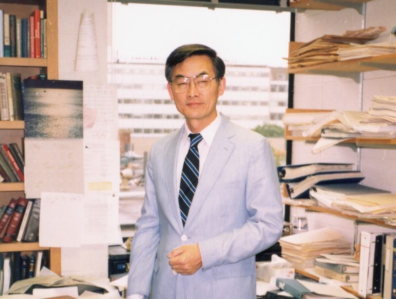 Color photo of C.C. Mei in middle age, standing in his office, with bookcases and a window