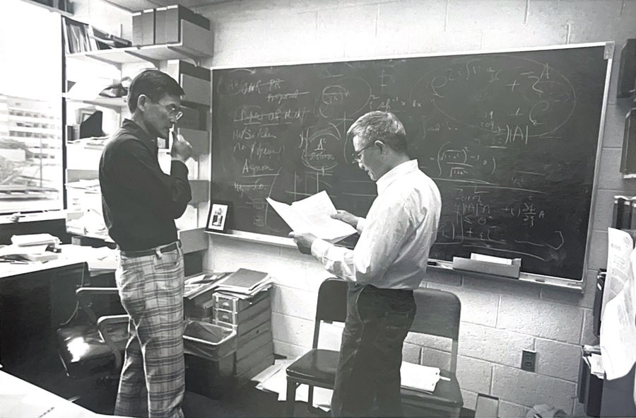 Grayscale photo of C.C. Mei holding a paper and talking to a student in a classroom with a blackboard behind them.