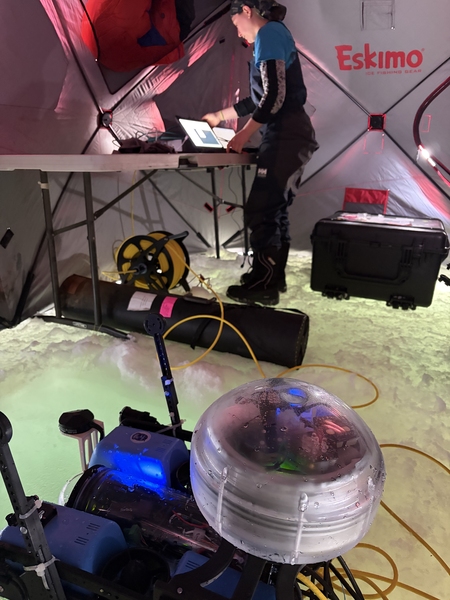 A remotely operated vehicle sits in a tent atop lagoon ice. In the background, Ella Wawrzynek works on a computer.