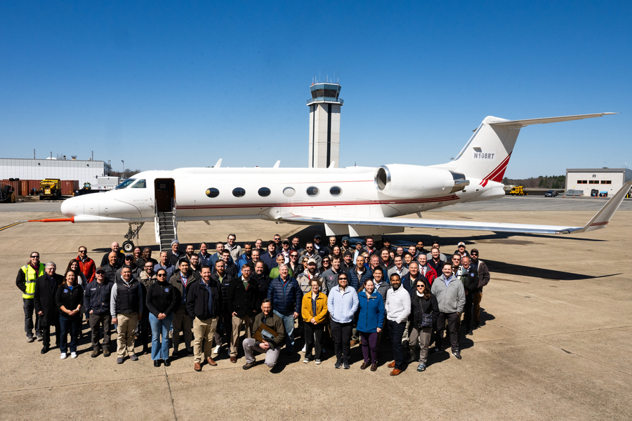 A large group of people in spring outerwear gathers before a plane on an airport tarmac