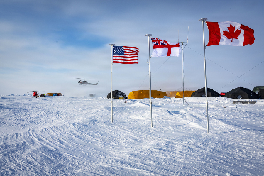 On a sunny day, multiple tents sit on Arctic sea ice, with flags of the U.S., U.K., and Canada in foreground