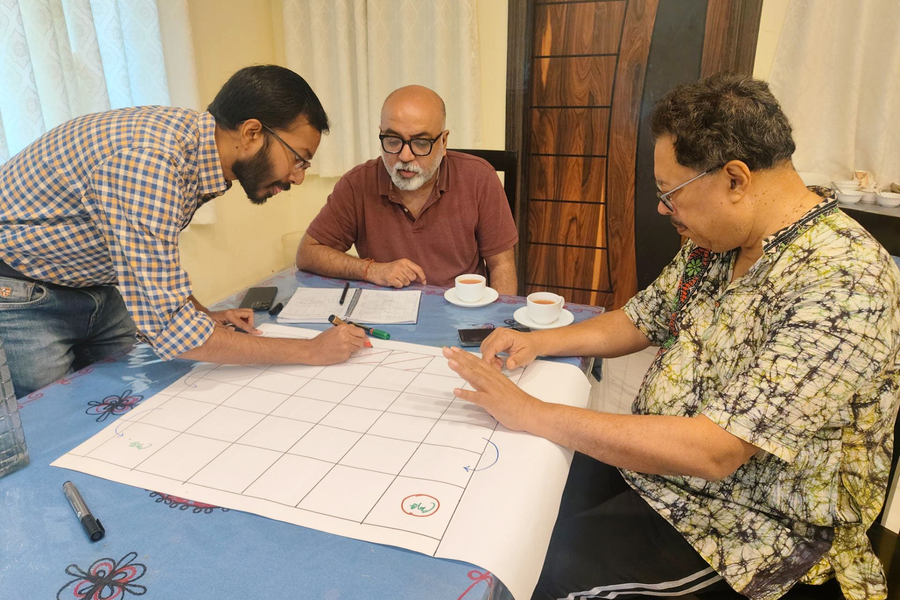 Three people sit at a table with markers, drawing symbols onto a large piece of paper lined with a grid.