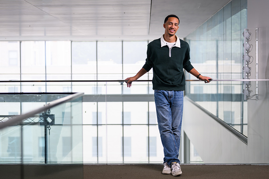 Kayode Dada leans against a glass railing with glass windows behind him.