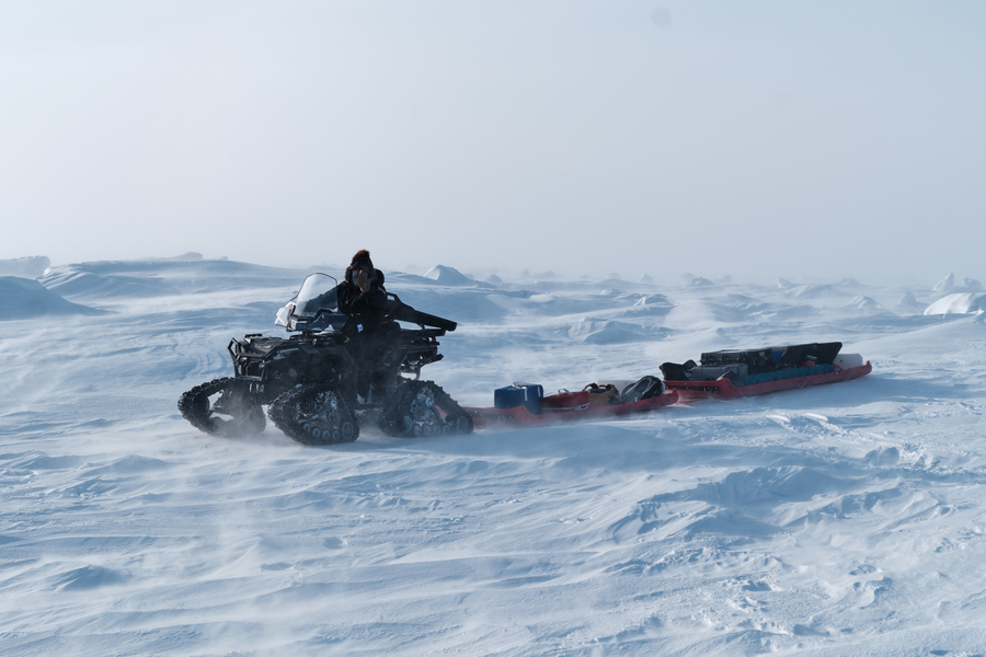 A person drives 4x4 vehicle with tracks, like a souped-up snowmobile, pulling a sled with equipment