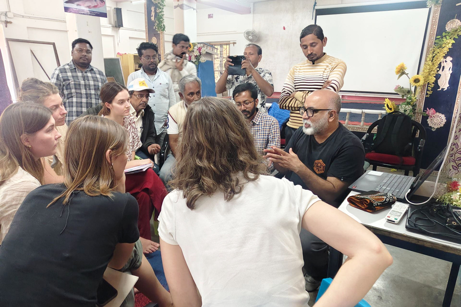 14 people, some holding notebooks, sit or stand in a collective huddle indoors.