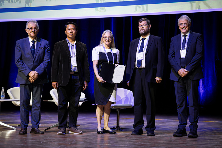 Anthea Coster poses, holding an award certificate on a stage, with four men at her side
