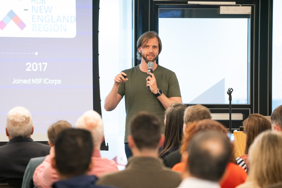 Ross Finman speaks to an audience, standing in front of a large classroom with a slide projected behind him.