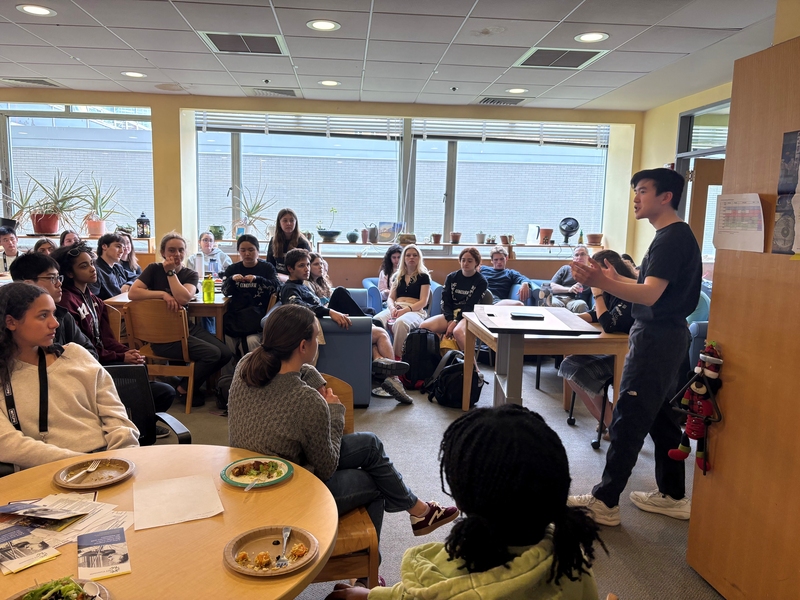 A student stands and speaks in a room while a couple dozen others, seated at tables and couches, listen