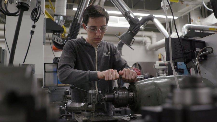 A young man wearing a hoodie and safety glasses operates a piece of heavy machinery