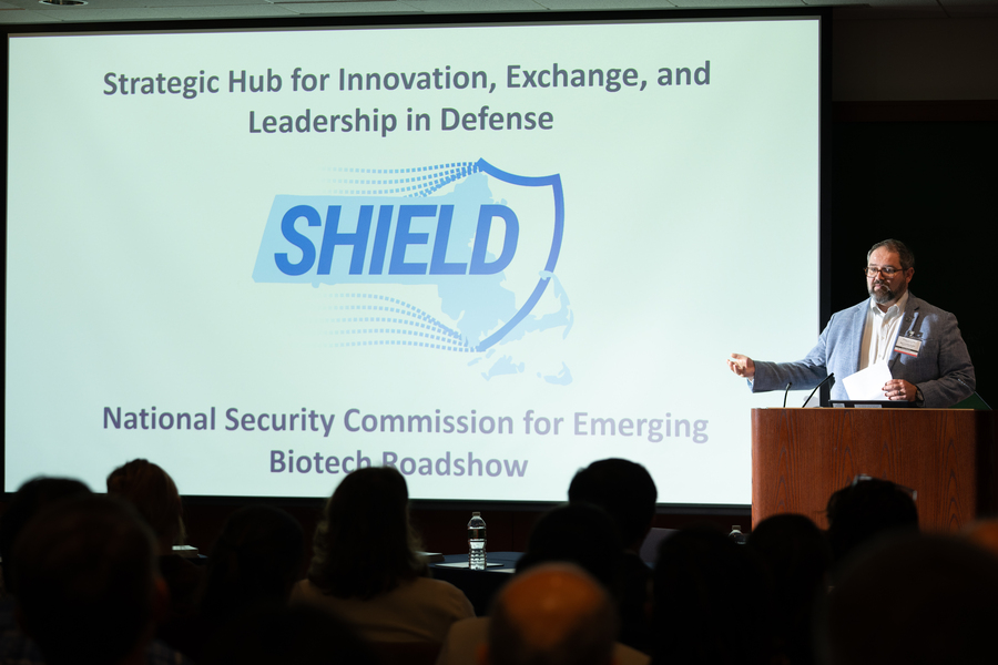 Ben Linville-Engler stands at a lectern in front of a screen displaying "SHIELD" and talks to a seated crowd.