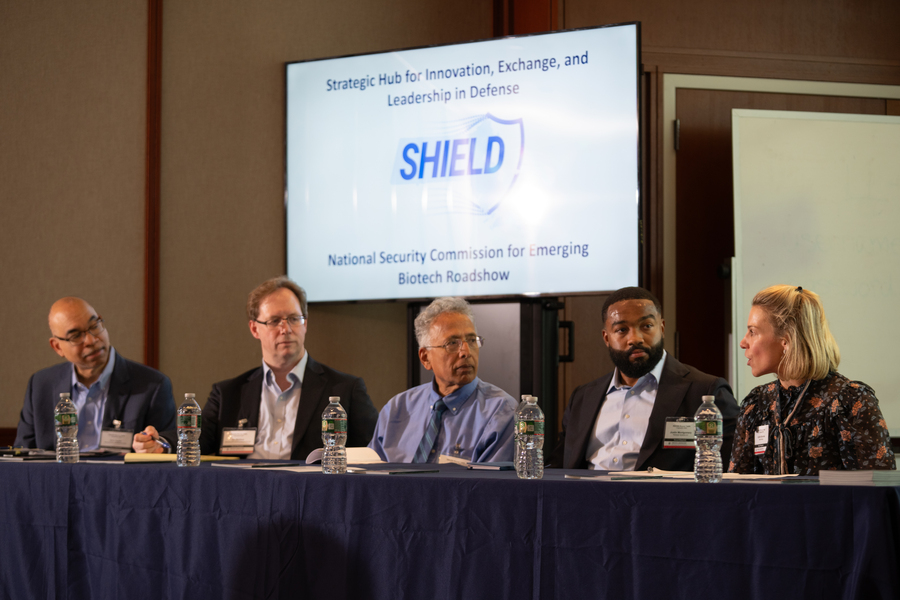 Five panelists sit at a table in discussion in front of a seated crowd. 