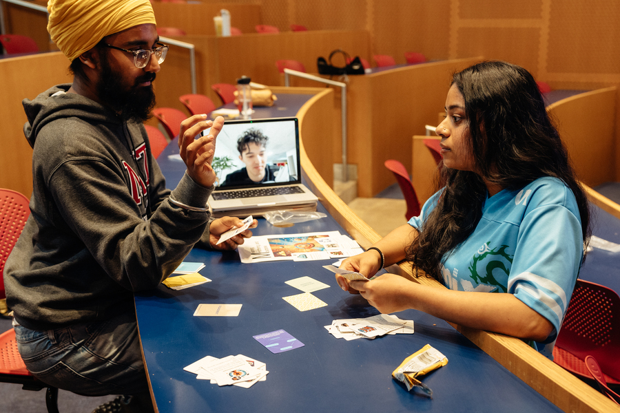 Amitoj Singh shows one of his collectible Intention card to a woman who sits across the desk from Amitoj, and another who is attending class via Zoom on an open laptop.