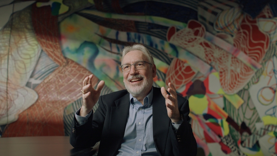 Duane Boning, seated in front of a colorful mural, gestures with both hands