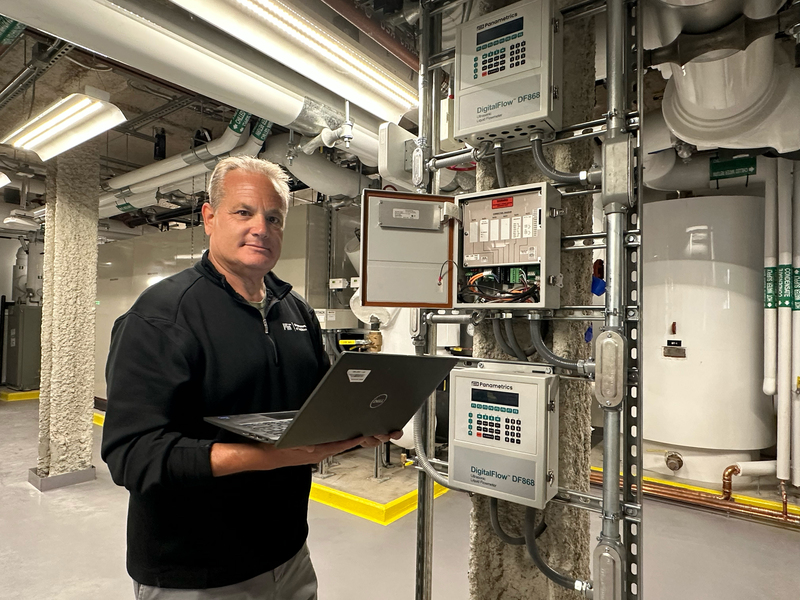 Fran Selvaggio holds an open laptop, standing beside equipment and pipes in a mechanical room.