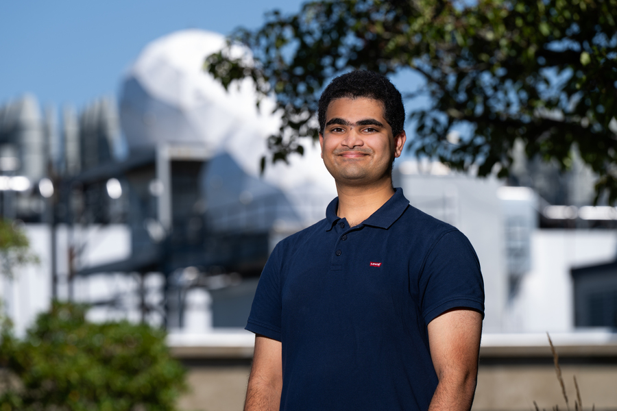 Vivek Jagadeesh portrait photo outside of Lincoln Laboratory, with radar antenna radomes in the background.