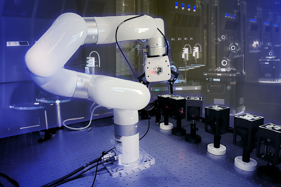 A white robotic arm lines up black boxes in an experiment.  A rendering of a futuristic robot lab is in the background.