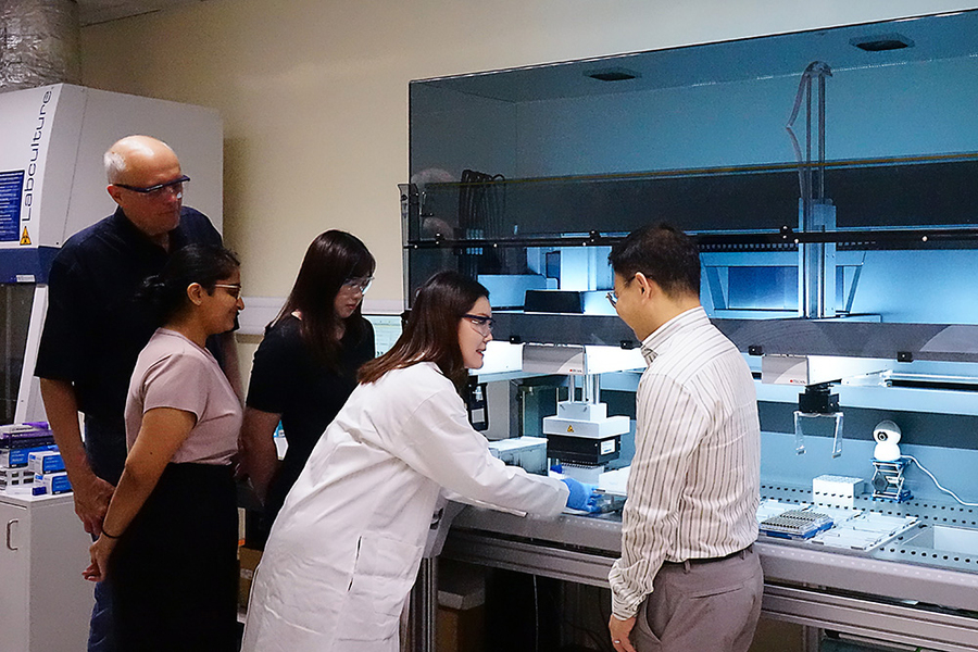 A woman in a lab coat operates equipment on a lab bench while four others look on