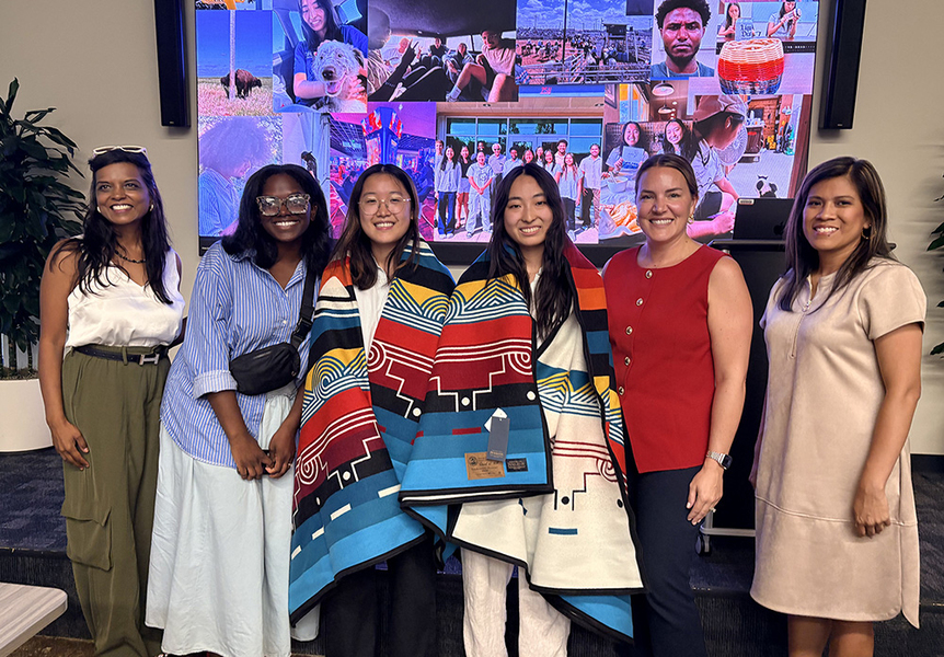 Six women pose standing in front of an electronic photo montage. Two in the center wear traditional Indigenous blankets.