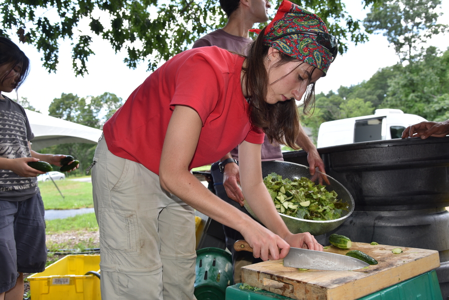 Aleks Banaś bends over a cutting board, slicing a cucumber with a chef’s knife.