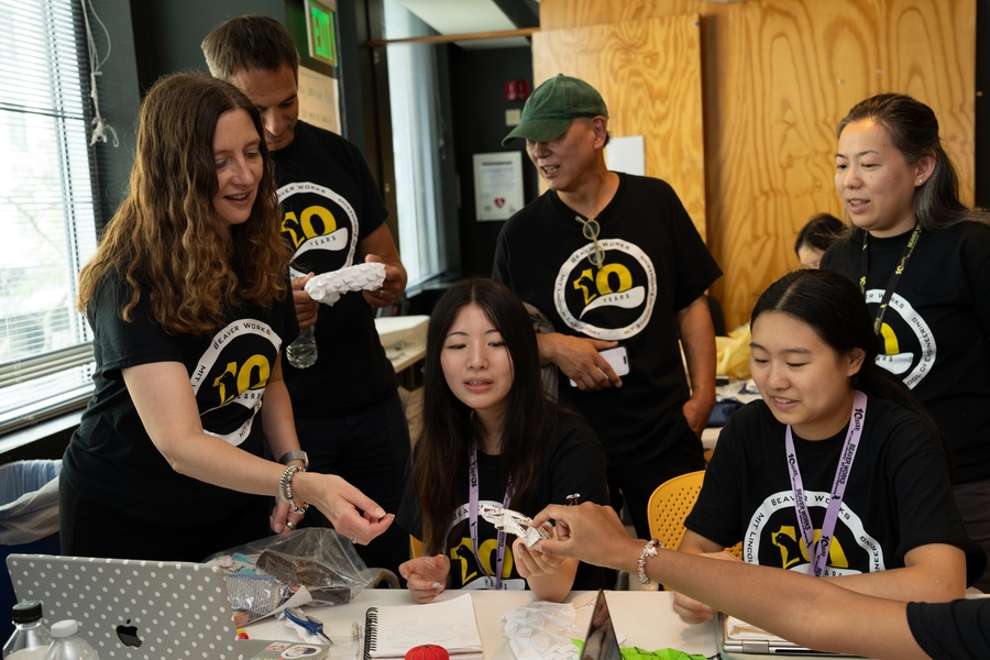 Melissa Choi interacts with students at a table filled with project items. All wear a BWSI black shirt with logo. 
