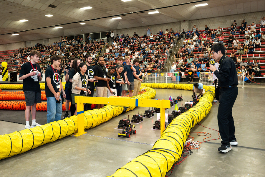 13 race participants, most wearing matching T-shirts, stand behind yellow tubing that denotes a racetrack inside an indoor arena. The racecar vehicle entries are gathered at a starting point while spectators watch on bleachers in the background.  