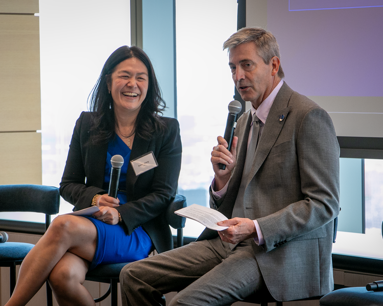Evelyn Wang sitting alongside Tom Taylor, holding microphones in front of a display screen.