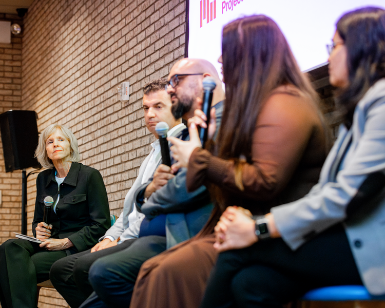 Elisabeth Reynolds sits at the left of a panel of five people, listening to one of them make remarks.