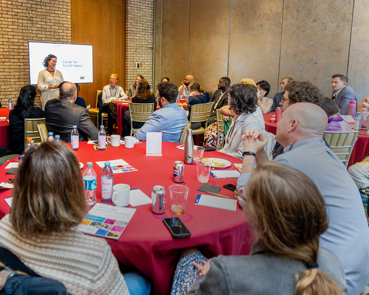 An audience sits at round tables in the foreground of small room, listening to a speaker at the far side standing at a lectern.