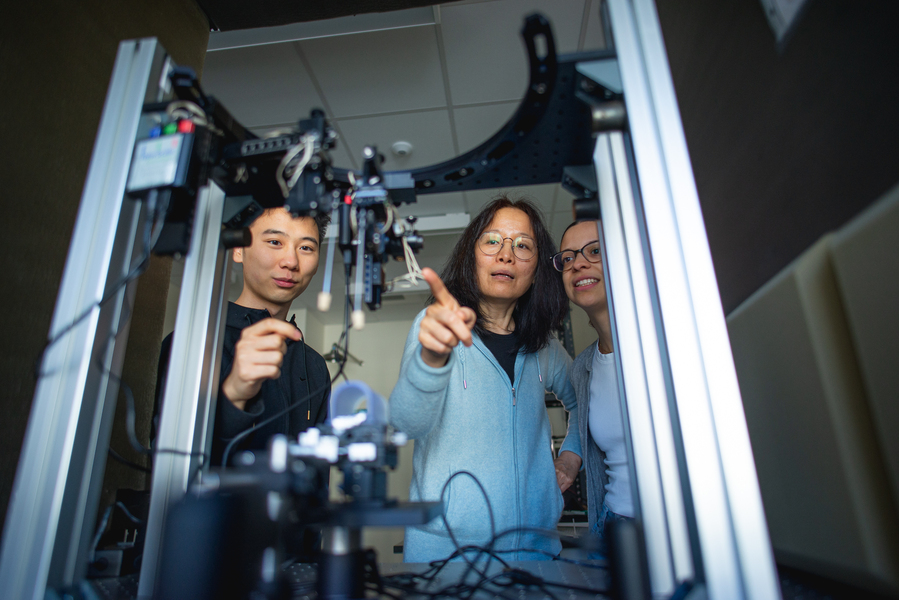 Fan Wang stands between two students, pointing to some lab hardware