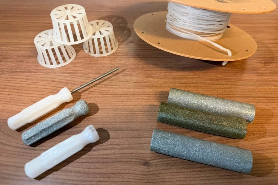 3D printed materials and tools on a wooden table: white spool holders, a large thread spool, plastic-handled awls, and glittery rods