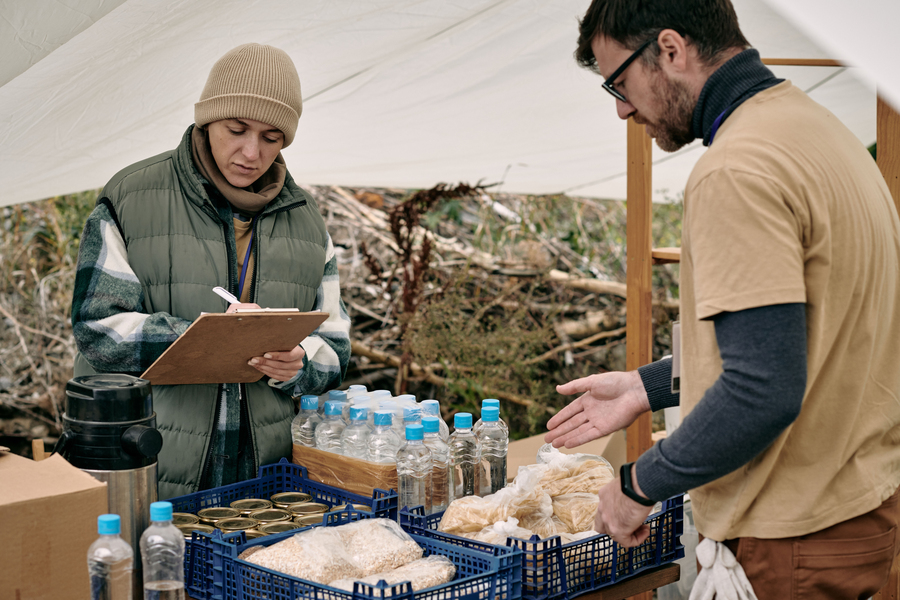 Two people stand outside under a tent. They are taking stock of supplies of bottled water, cans of food, and food packages in crates.