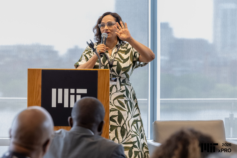 Prabha Jha speaks at a lectern in front of glass walls overlooking the Charles River