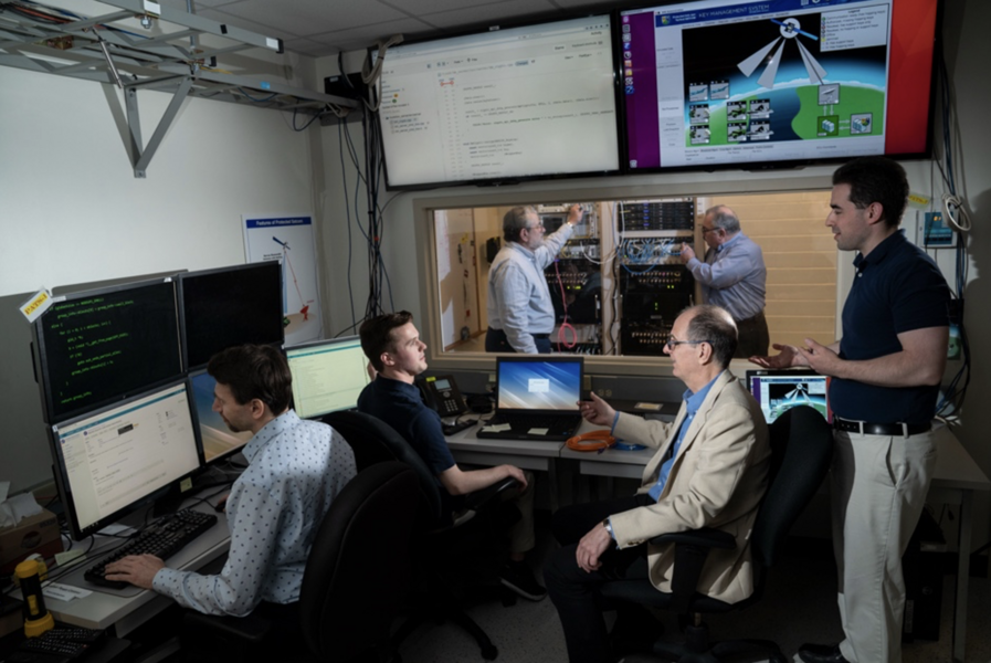 Six researchers are in a lab with computer screens, monitors, and two server racks of hardware 