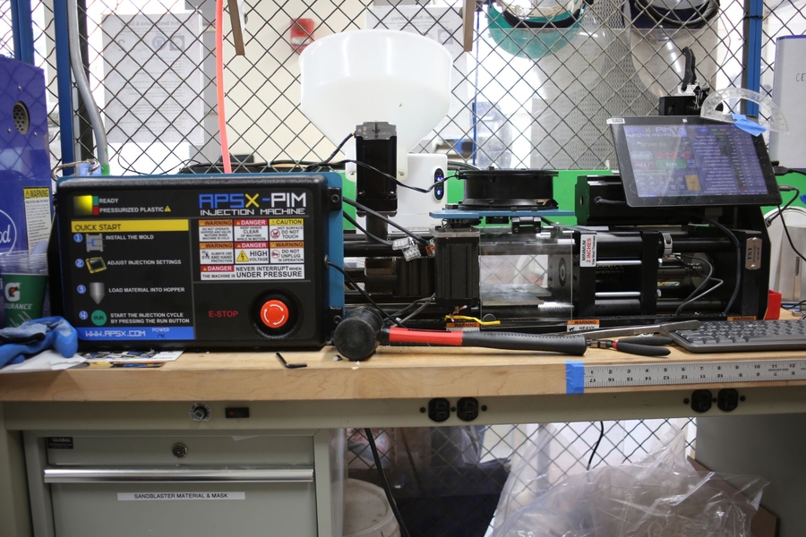 A 3D-printing workstation with a filament spool, printer, touchscreen, and control unit on a cluttered bench in a wire-mesh workshop