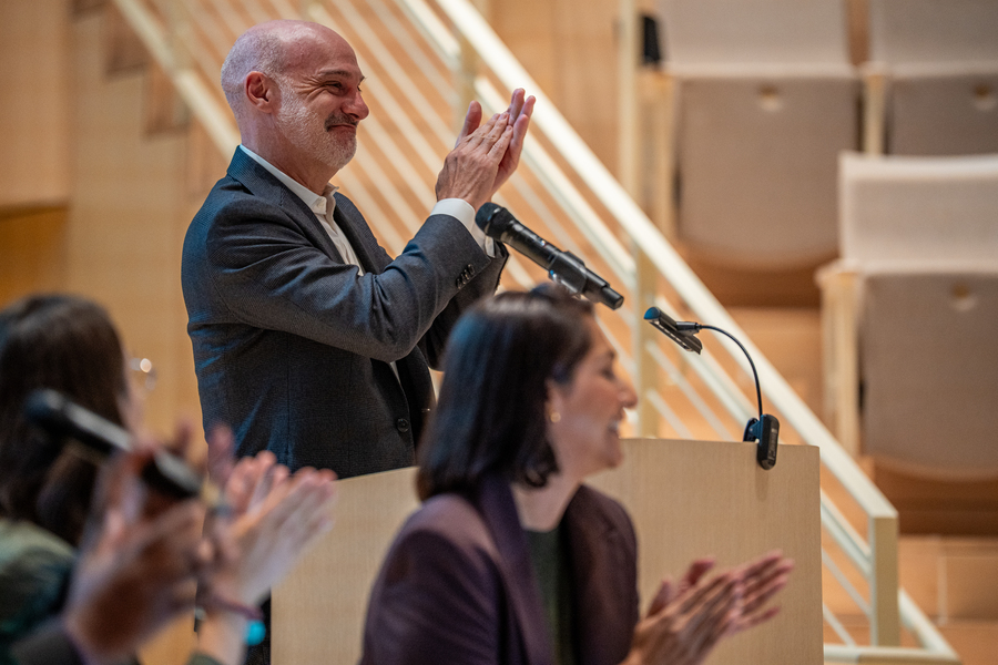 Agustin Rayo stands at a lectern, smiling and applauding something out of the frame