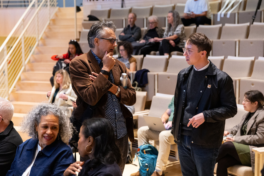 Two attendees in a sparsely populated auditorium discuss the 75th Anniversary Symposium