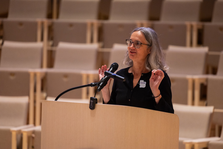 Heather Paxson speaks into a microphone at a lectern, with a bank of empty seats behind her