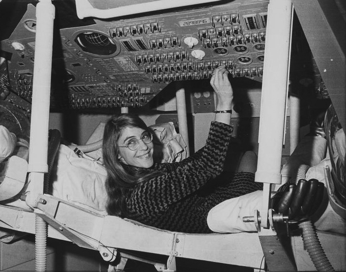 Greyscale photo of a young Margaret Hamilton posing in a mockup of an Apollo capsule