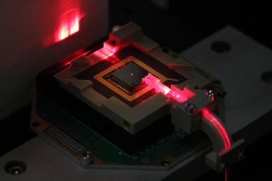 Red laser light from a fiber optic cable hits the side of a small square chip.