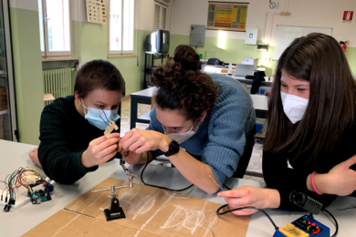 Photo of three students, all wearing facemasks and leaning on a lab table and holding simple electronic equipment
