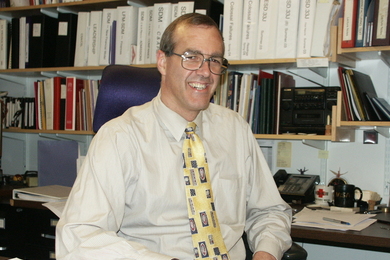 Man in a tan shirt, yellow tie, and glasses is smiling at his in front of a crowded book case 