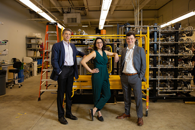 Photo of a woman standing between two men in a lab with lab equipment in the background.