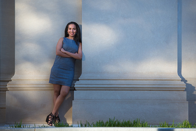 A young woman with long dark hair, wearing a blue dress and folding her arms, stands leaning against a stone facade