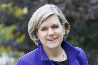 Portrait photo of Nancy L. Rose, wearing a purple coat and standing in front of a tree in summer